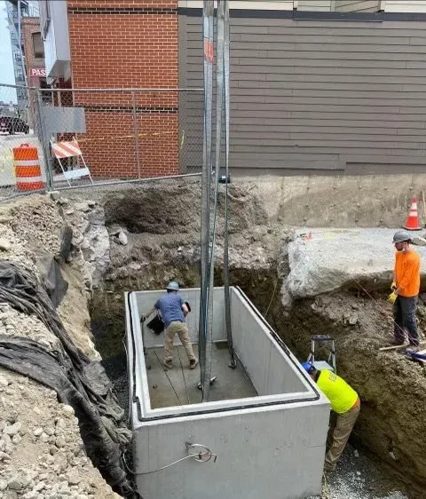 Construction workers are installing a large underground utility vault at a construction site, surrounded by dirt and construction equipment.
