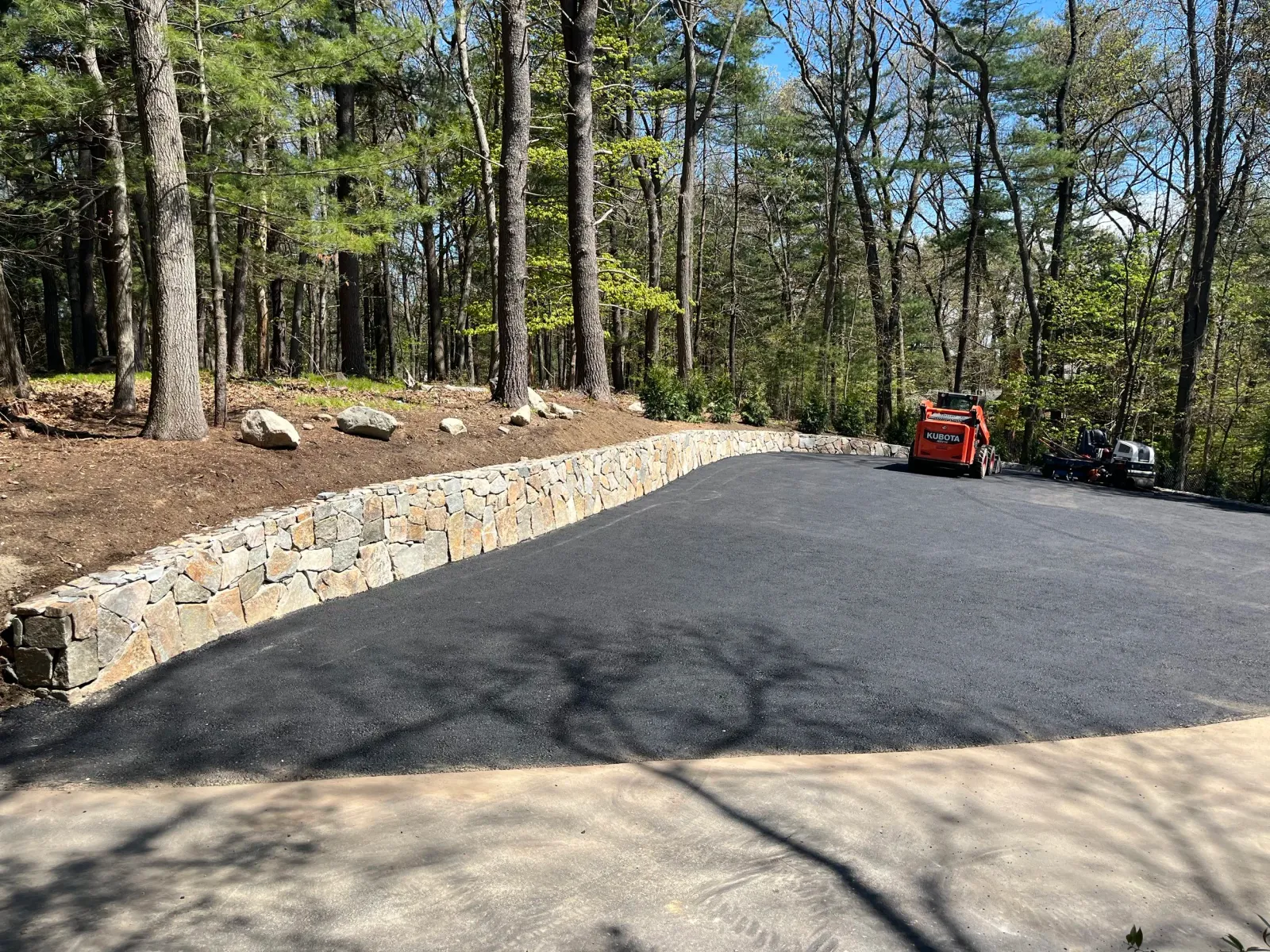 A newly paved blacktop driveway with a stone retaining wall on the left side, surrounded by trees in a wooded area, with construction equipment parked at the end of the driveway.