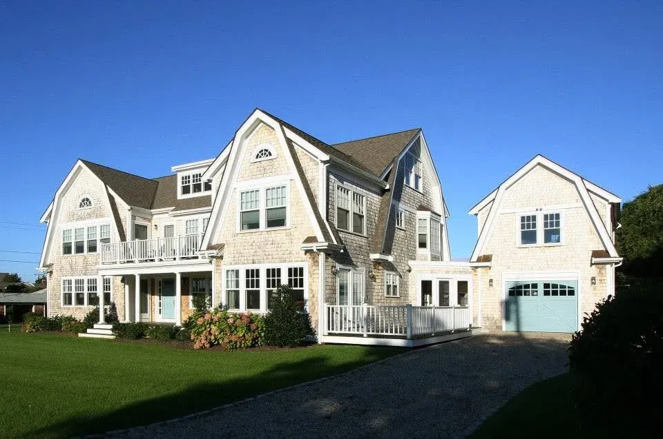 Large house with multiple stories, light-colored siding, and a detached garage, set against a bright blue sky, with a well-maintained green lawn and shrubs in the front yard.