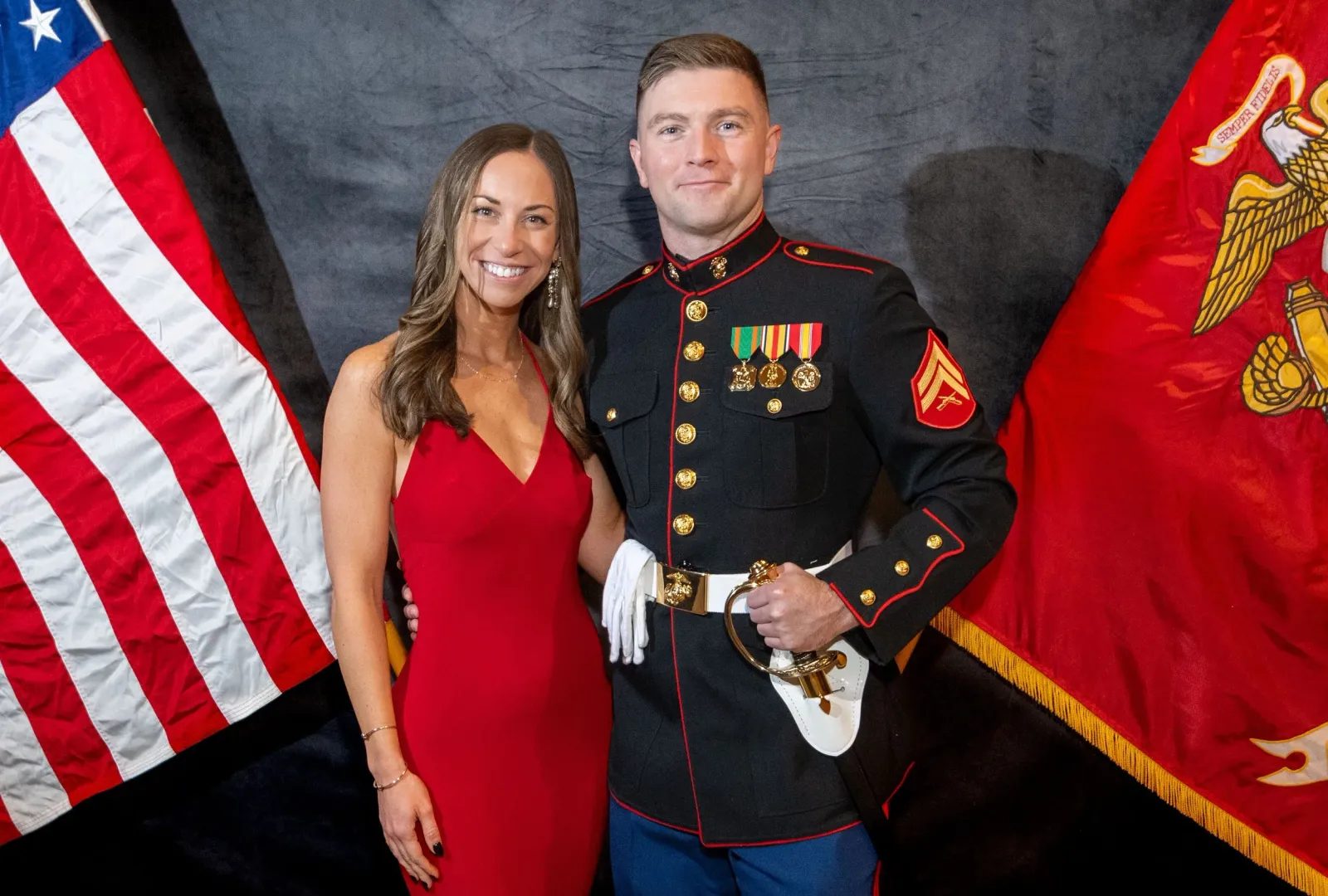 A woman in a red dress and a man in a U.S. Marine uniform standing together in front of American and Marine Corps flags.
