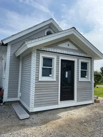 Small gray house with white trim and black front door on a gravel lot