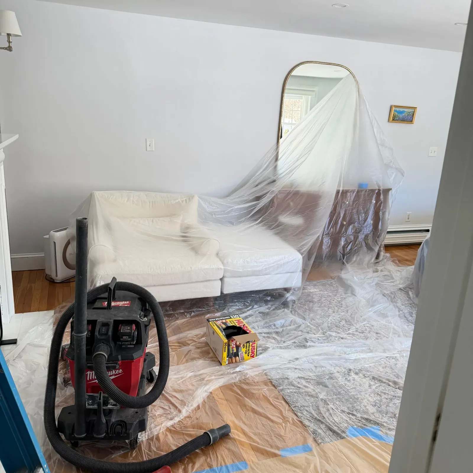 Living room under renovation, with sofa covered in plastic and a vacuum on the floor.