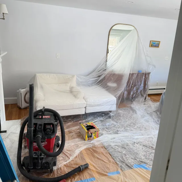 Living room under renovation, with sofa covered in plastic and a vacuum on the floor.