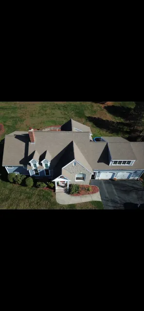 Aerial view of a suburban house with a brick chimney, gray roof, front porch with plants, and attached garage. Surrounding lawn and trees are visible.