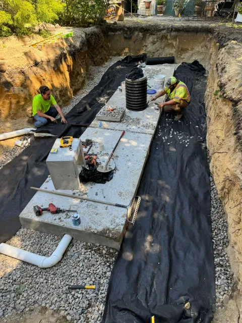 Two workers installing a septic system in a trench. Concrete tanks sit on gravel; black fabric lines the pit.