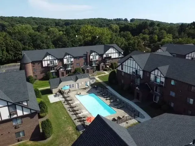 Aerial view of a residential apartment complex with a swimming pool, surrounded by green trees and hills in the background.