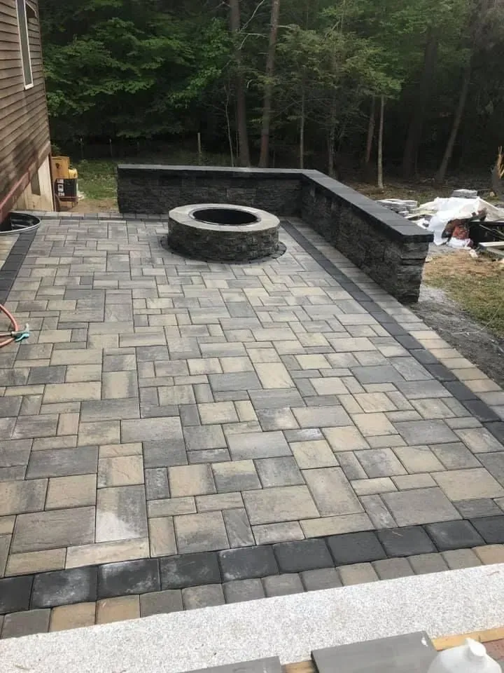 Stone patio with circular fire pit, bordered by a dark retaining wall and woods beyond.