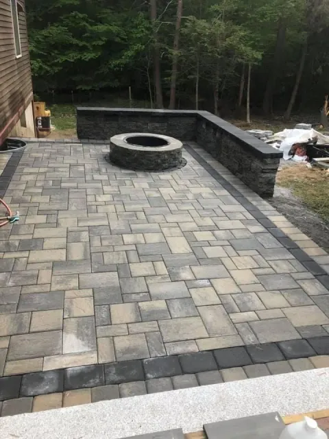 Stone patio with circular fire pit, bordered by a dark retaining wall and woods beyond.