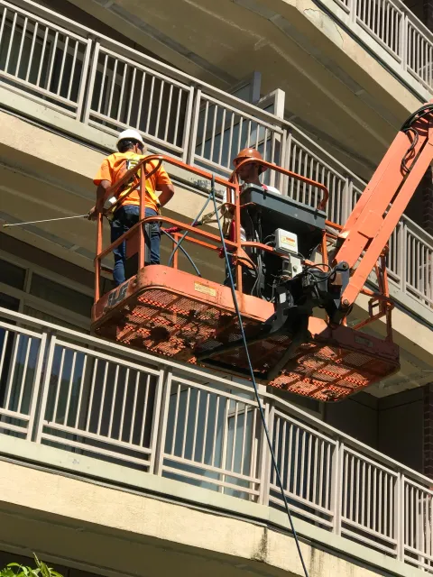 Two construction workers on an orange aerial lift performing maintenance on a multi-story building's balcony railing