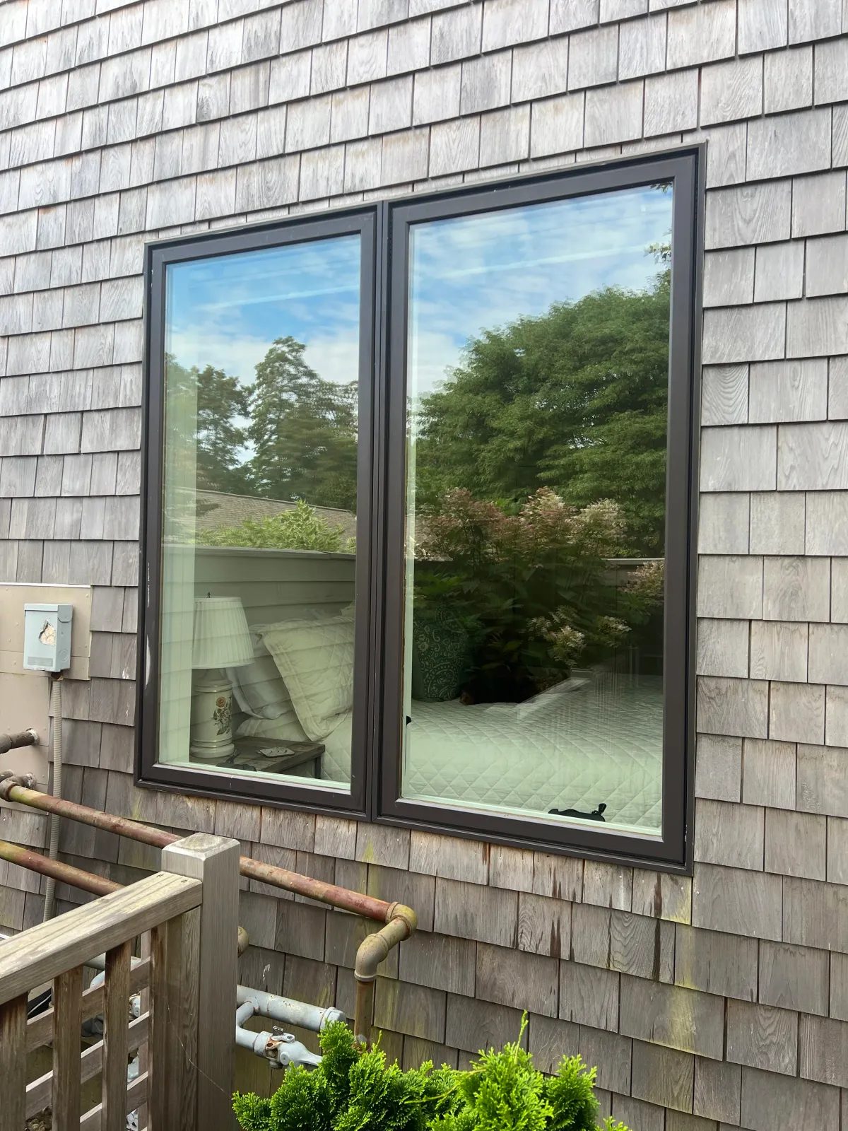 A window on a weathered wooden exterior wall reflecting trees and a cloudy sky, with a view of a bedroom interior featuring a bed, pillows, a nightstand with a lamp, and some plants outside.