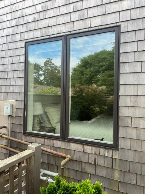 A window on a weathered wooden exterior wall reflecting trees and a cloudy sky, with a view of a bedroom interior featuring a bed, pillows, a nightstand with a lamp, and some plants outside.