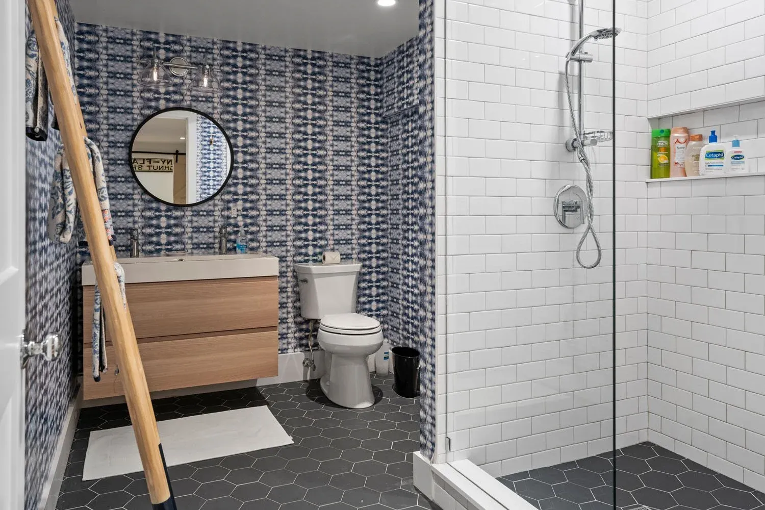 Modern bathroom with a patterned blue wallpapered wall, a white toilet, a wooden vanity with a round mirror, a towel ladder, and a walk-in shower with white subway tiles and shelves holding toiletries.
