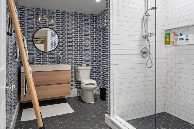 Modern bathroom with a patterned blue wallpapered wall, a white toilet, a wooden vanity with a round mirror, a towel ladder, and a walk-in shower with white subway tiles and shelves holding toiletries.