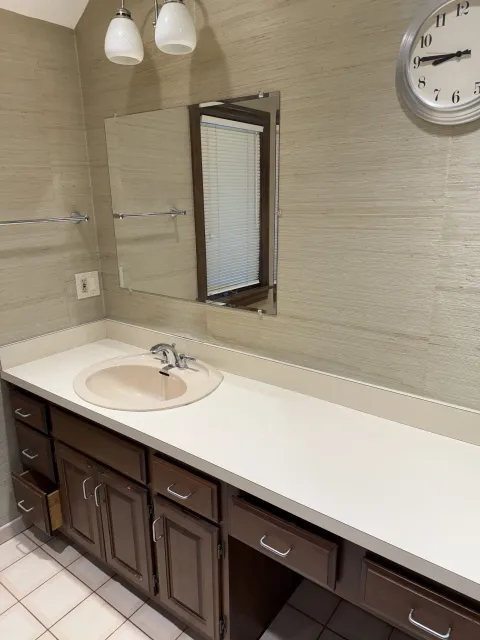 Bathroom with a vanity, mirror, clock, and two light fixtures. Beige walls, white countertop, dark cabinets.