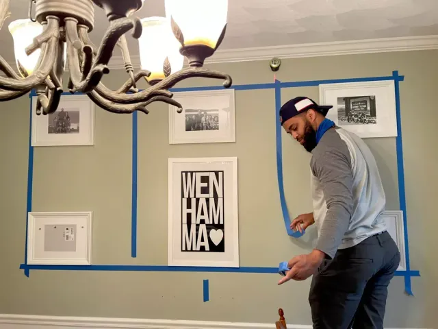 Person taping blue painter’s tape on a wall with framed photos and a chandelier overhead