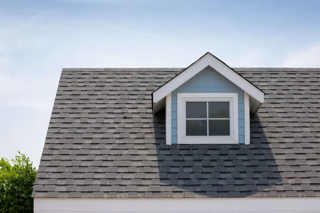 Gray shingled roof with dormer window and blue siding under a clear sky