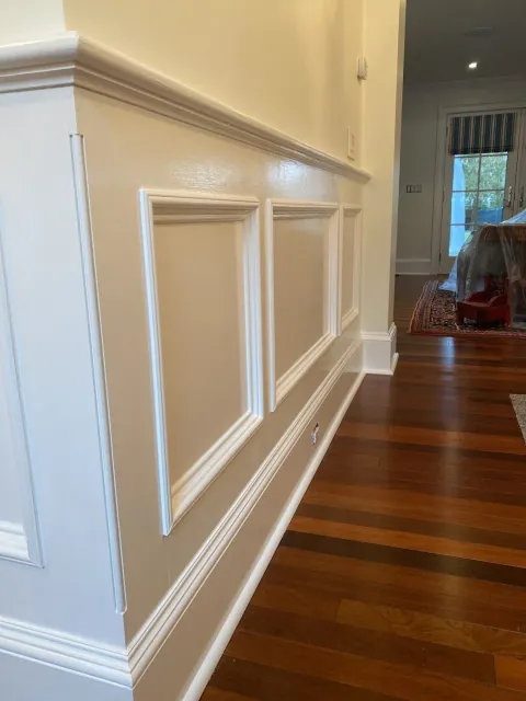 A wall with white wainscoting and square recessed panels. The wall is painted a light beige color. Hardwood flooring and a glimpse of another room are visible.