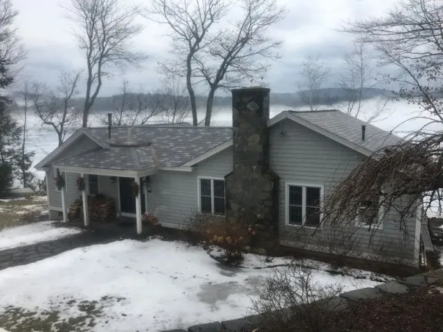 A gray house with a stone chimney sits on a snowy bank overlooking a lake on a cloudy day.