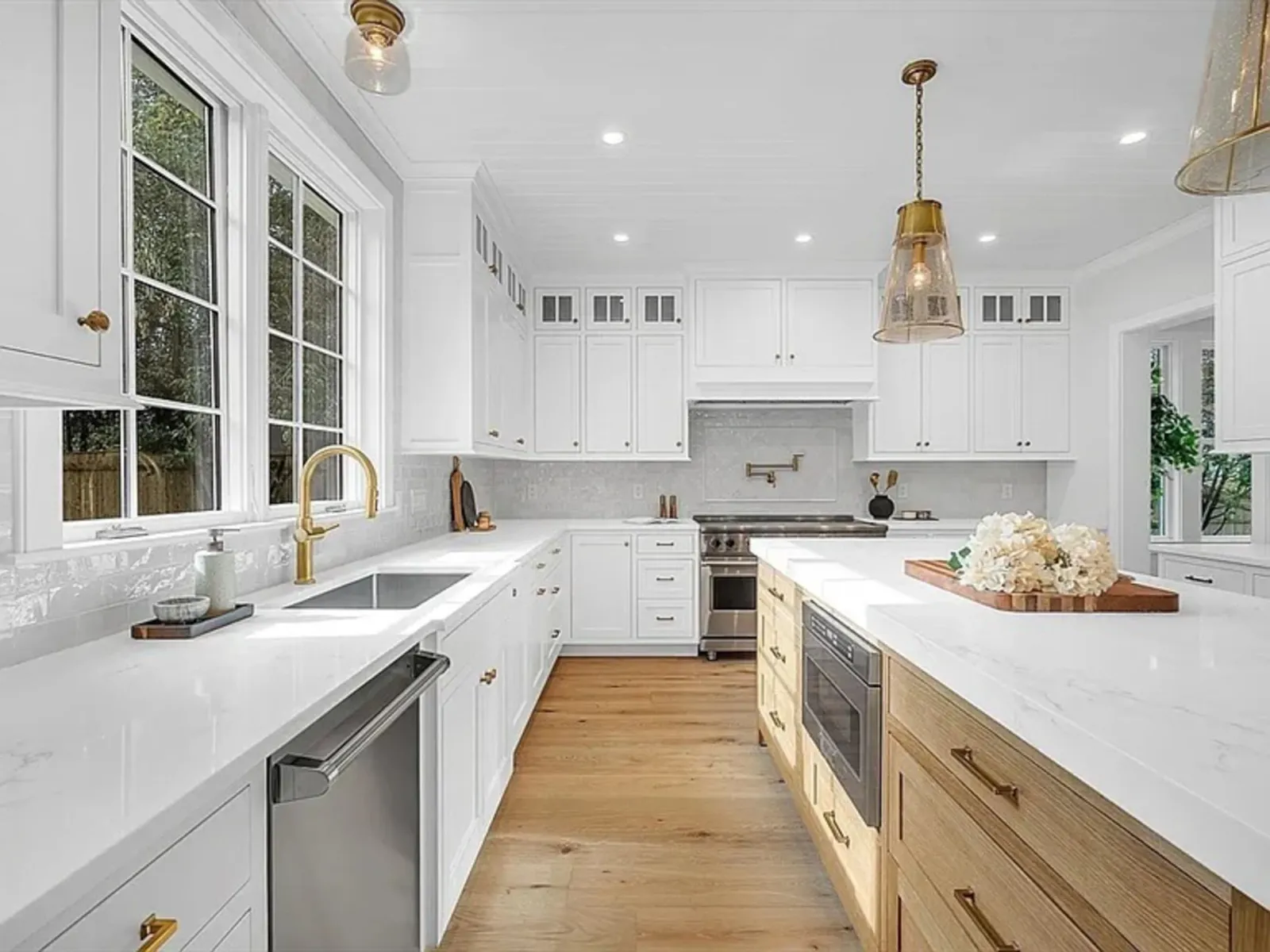 Hardwood floor installation in a Plymouth kitchen with white cabinets, gold fixtures, large windows, a marble island with flowers, and mixed wood and white countertops.
