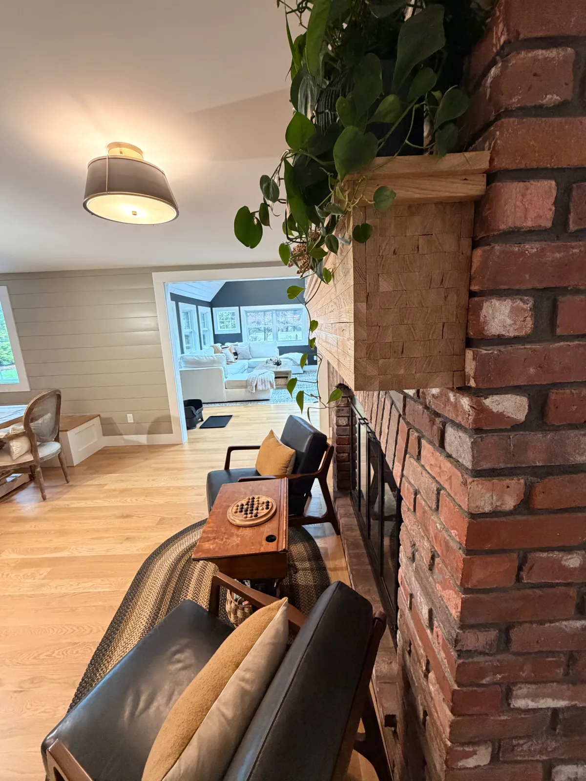 Living room with a brick fireplace, a wooden chair with cushions, black armchairs, and a dining table. Visible through an open doorway is a bright room with large windows and a white sofa.