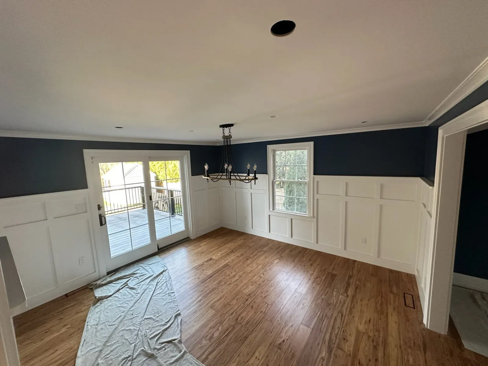 Empty living room with hardwood floors, dark blue walls with white wainscoting, a chandelier, and sliding glass doors leading to a deck.