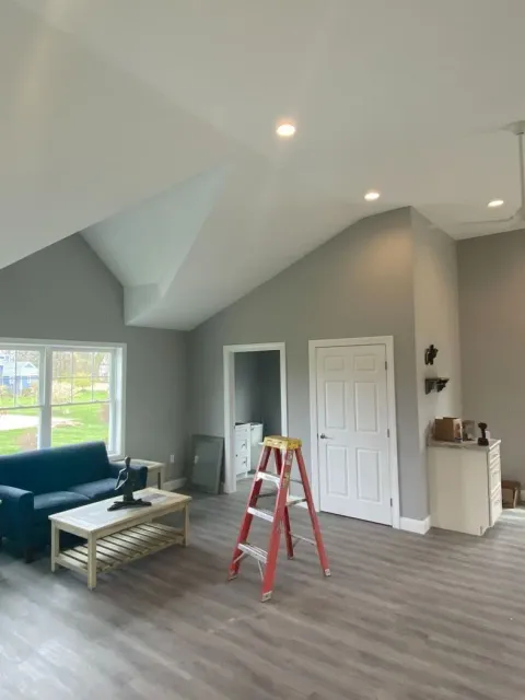 Spacious gray living room with vaulted ceiling, blue sofa, ladder, and open doorway.