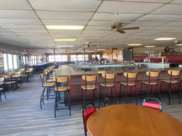 Empty restaurant or bar with wooden chairs and tables, a long counter with bar stools, ceiling fans, and large windows letting in natural light.