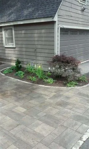 Driveway beside a gray house with a small landscaped garden bed and paver stone paving.