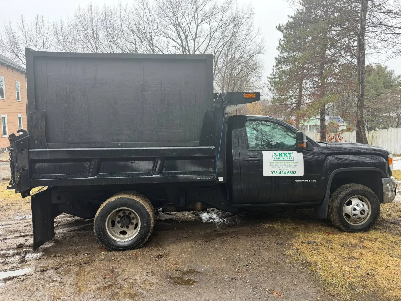 Black dump truck parked on muddy ground beside a building and leafless trees