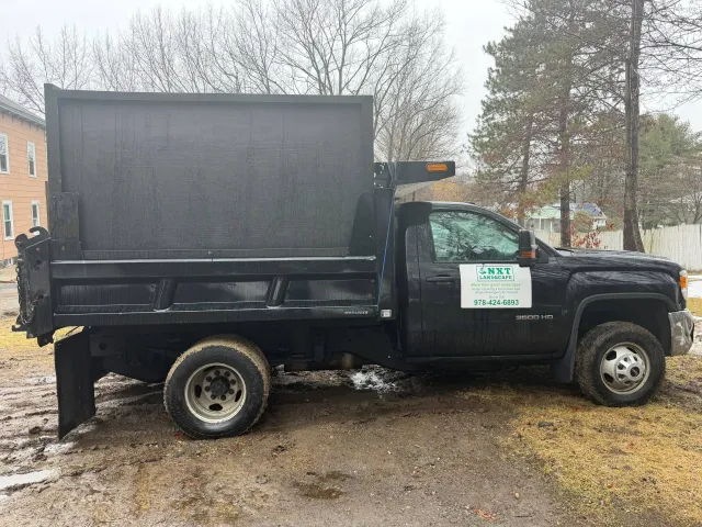 Black dump truck parked on muddy ground beside a building and leafless trees