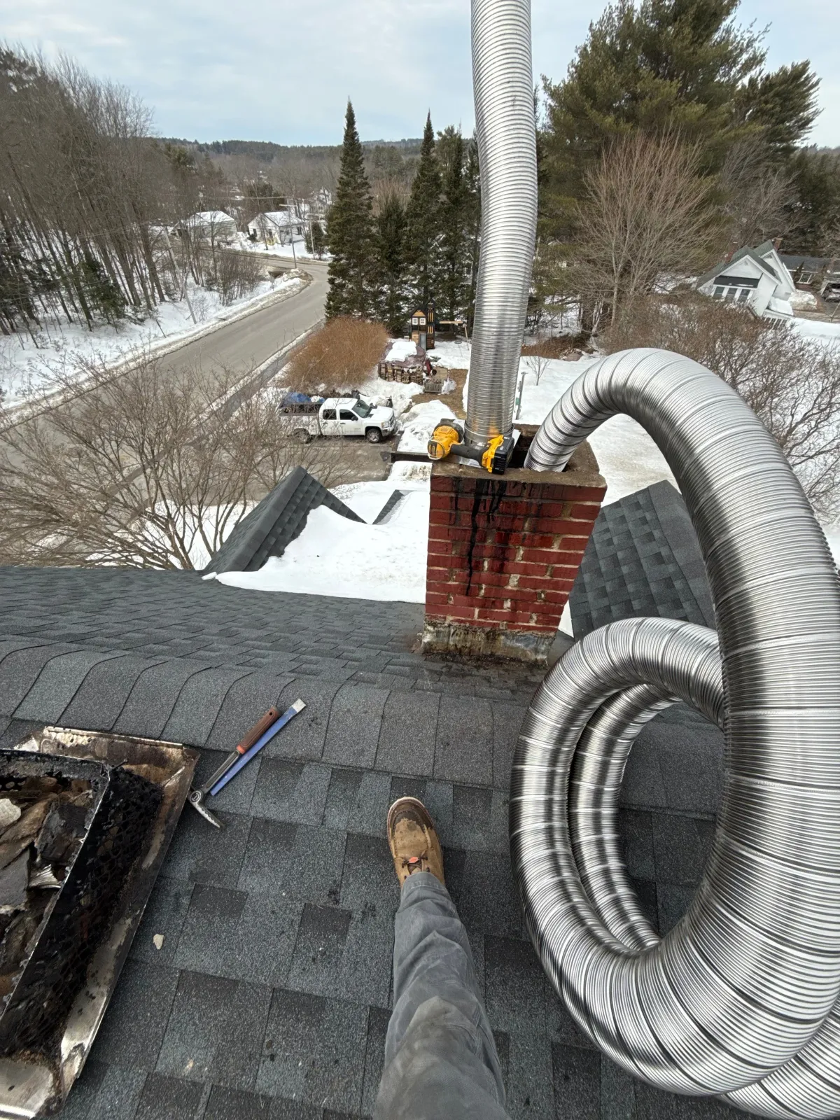 View from a rooftop with chrome chimneys, a brick chimney, and tools, overlooking a snow-covered neighborhood street with trees, trucks, and houses.
