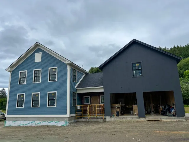 A house under construction with a house finished in blue siding and a garage finished with black siding. The house has six windows, and the garage is open with construction supplies and furniture inside.