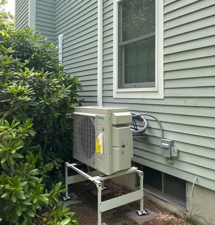 Outdoor HVAC unit beside a gray house with green siding, near bushes and a window.
