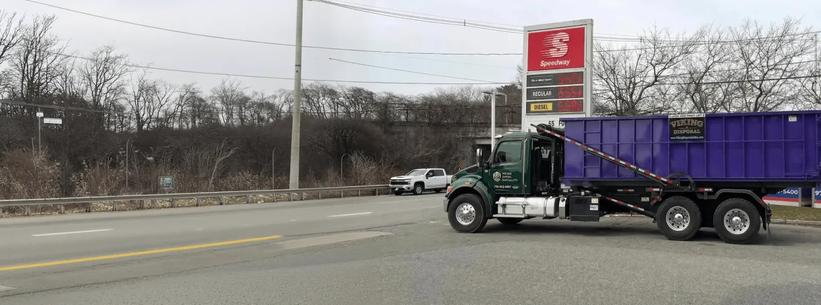 Green roll-off truck carrying a purple disposal container parked beside a roadside gas station sign on an overcast day.