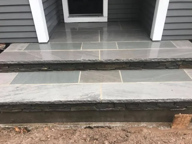 Close-up view of stone steps leading to a house with gray siding and a tiny porch. The steps are unfinished with visible dirt at the bottom.