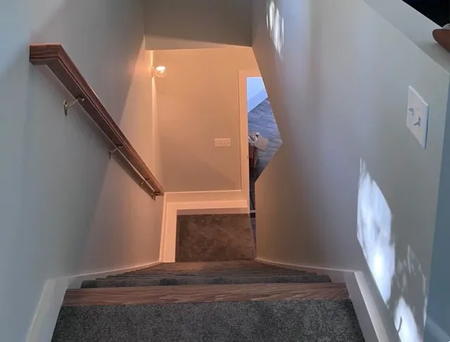 A downward view of a staircase with gray carpeted steps, a wooden handrail on the left wall, and a light switch on the right wall. At the bottom of the stairs, there is a small landing area with a door open to a room.
