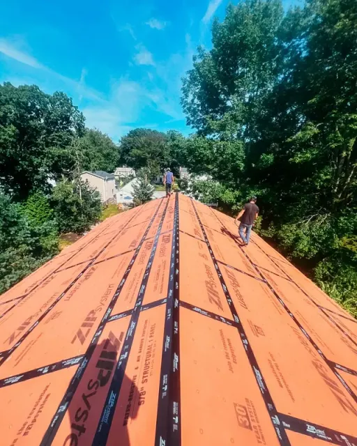 Two workers on an orange roof under construction, surrounded by trees and blue sky