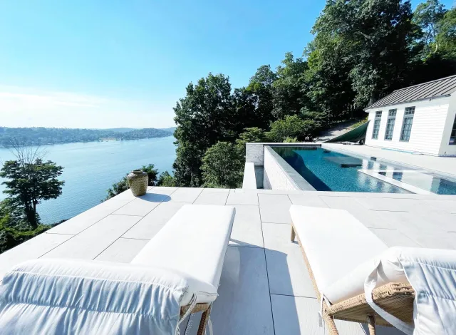 Luxury outdoor infinity pool overlooking a large body of water, with two white lounge chairs on a white tiled patio, surrounded by lush green trees under a clear blue sky.
