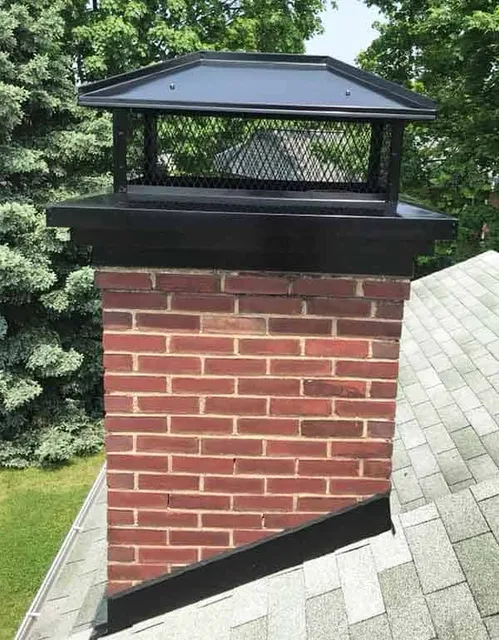 A red brick chimney with a black metal cap and mesh sides sits atop a shingled roof, surrounded by green trees in the background—ideal for Chimney Cleaning Providence County, RI homeowners.