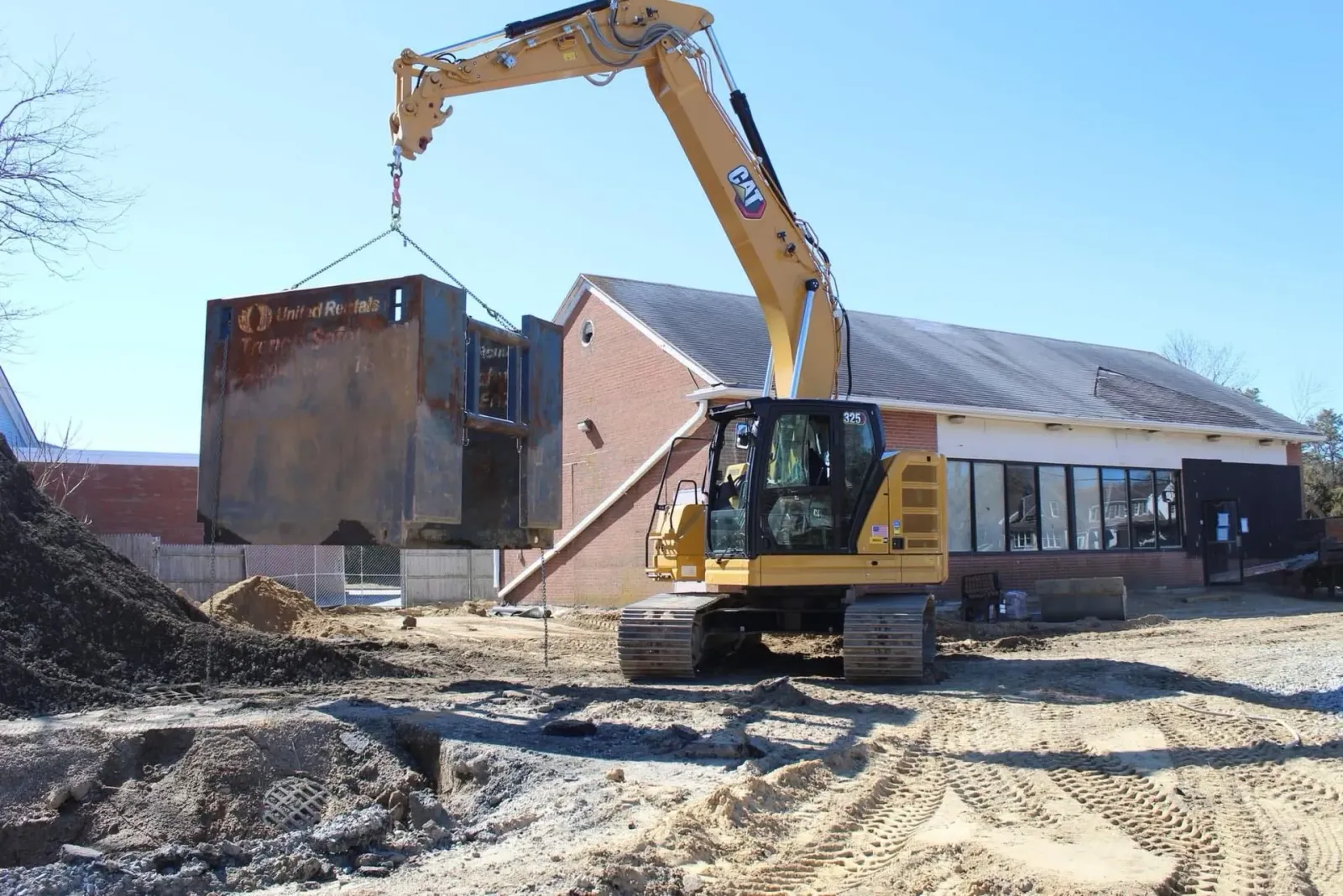 Yellow excavator lifting a large concrete block at a construction site near a brick building with a white roof.