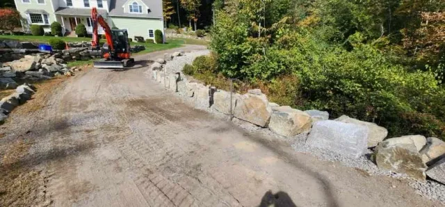 Excavator on dirt path near house with stone embankment and green foliage