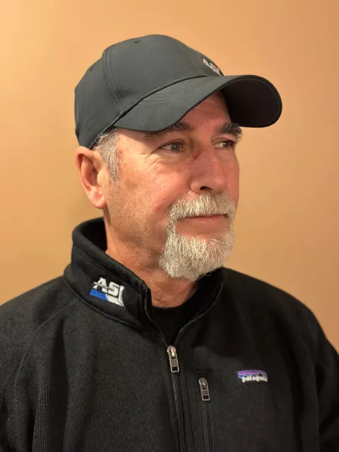 A middle-aged man with gray hair and a gray beard wearing a dark baseball cap and a black Patagonia jacket, looking to his left against a plain beige wall.