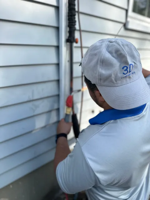 A man working outside on siding of a house, using a power tool near a pipe attached to the wall, wearing a white cap and white shirt with blue accents.