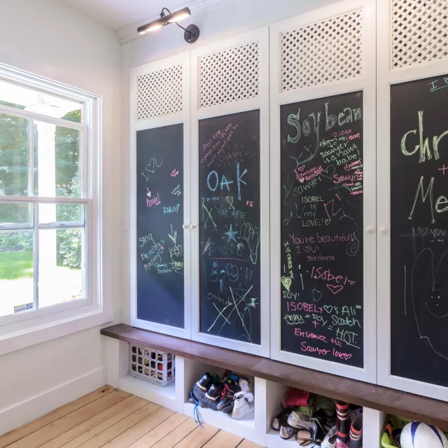 A corner of a room with a window showing trees outside, a black chalkboard wall with colorful handwritten messages and drawings, a ceiling light, a wooden floor, and shelves filled with shoes and sports equipment.