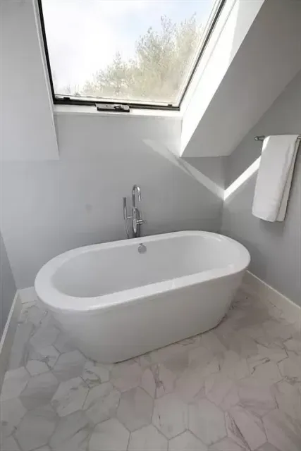 Freestanding white bathtub under a sloped skylight in a bathroom with light grey walls and hexagonal marble floor tiles.