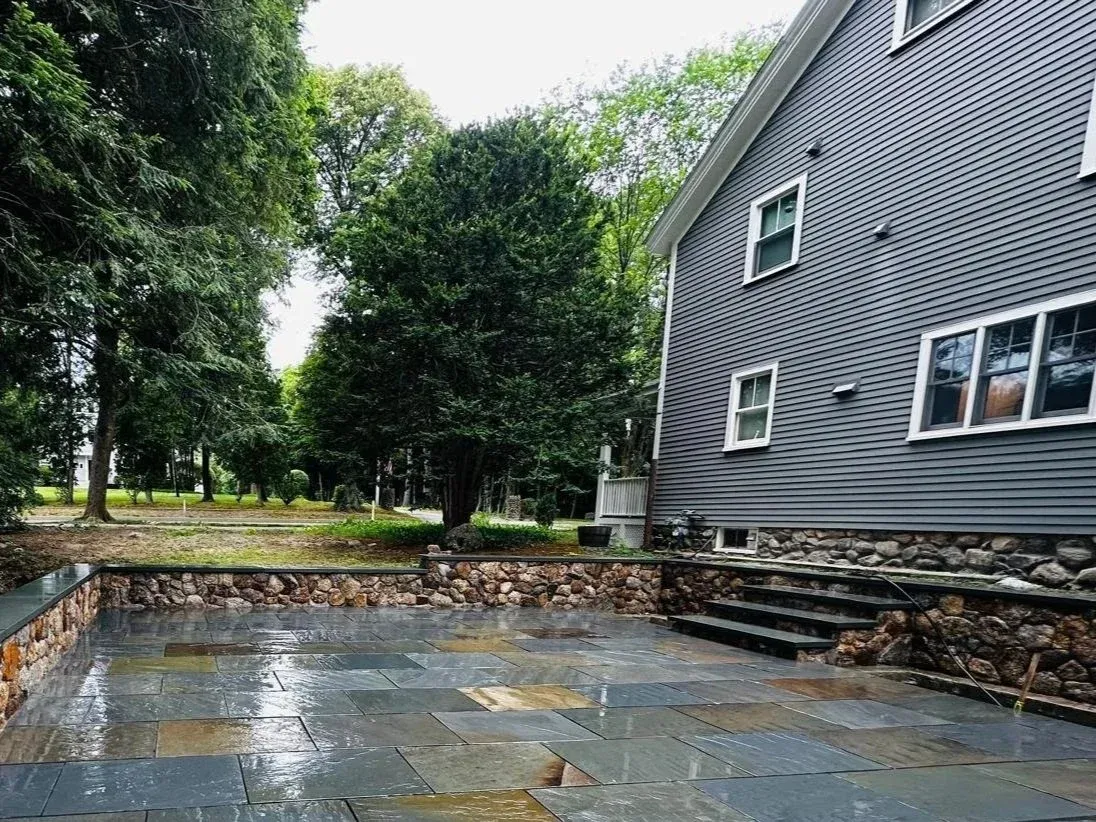 A wet slate patio next to a gray house with white window frames, stone foundation, and steps leading to the yard, surrounded by green trees.