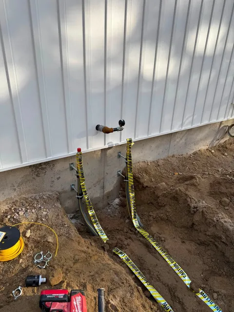 Two yellow tie-down straps hanging from a wall-mounted bracket over a dirt trench beside a white fence