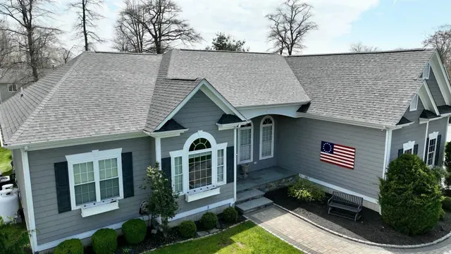 A grey suburban house with a shingled roof, a paved walkway, landscaping, and an American flag displayed on the wall.