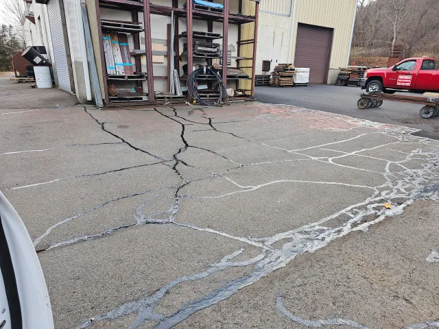 A cracked asphalt parking lot surface with visible sealant lines, near a warehouse building with metal storage racks.