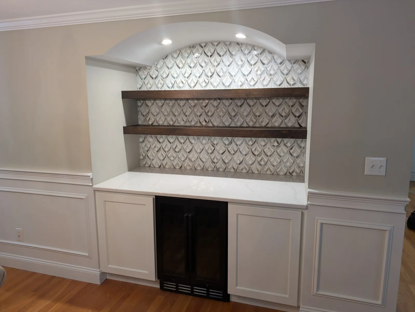 Interior view of a kitchen or living space with a built-in shelving unit featuring two dark wooden shelves and a patterned marble-like background, white cabinetry, and hardwood flooring.